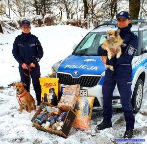 Dwie umundurowane policjantki z dwoma psami stoją obok oznakowanego radiowozu . Przed radiowozem znajduje się karma dla zwierząt w tle śnieg