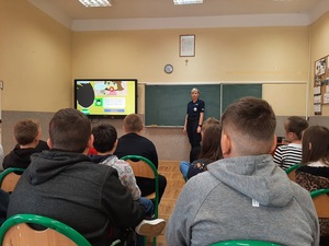 spotkanie policjanta z uczniami, klasa uczniowie w ławkach w tle policjantka umundurowana