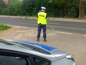 radiowóz przy drodze, dzień, umundurowany policjant w żółtej kamizelce stoi tyłem w ręku trzyma fotoradar skierowany w stronę ulicy