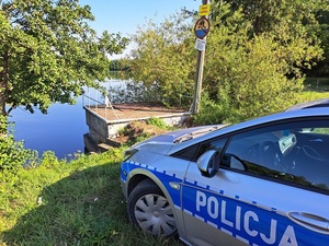 zaparkowany radiowóz oznakowany na drodze gruntowej w okolicy akwenu wodnego