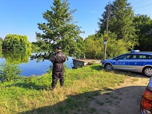 zaparkowany radiowóz oraz osoba ubrana na ciemno na tle akwenu wodnego
