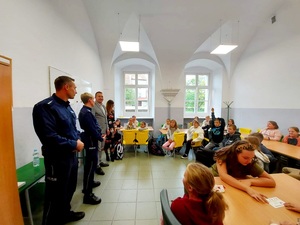 policjantka i policjant stoją, dzieci w wieku szkolnym siedzą  na krzesłach w klasie