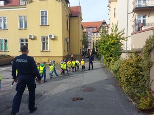policjant stoi, dzieci idą w parach w kamizelkach odblaskowych