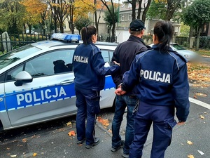 dwie umundurowane policjantki i pomiędzy nimi mężczyzna w kajdankach na tel oznakowanego radiowozu