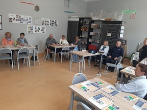 seniorzy siedzą przy stołach w pomieszczeniu, policjant umundurowany siedzi przy komputerze