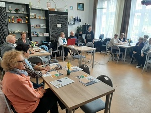 seniorzy siedzą przy stołach w pomieszczeniu, policjant umundurowany siedzi przy komputerze