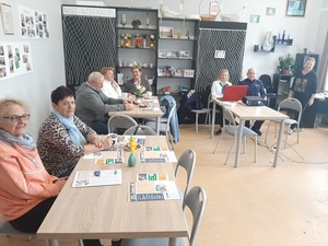 seniorzy siedzą przy stołach w pomieszczeniu, policjant umundurowany siedzi przy komputerze