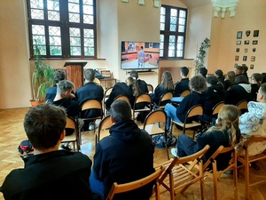 policjantka stoi przy ekranie telewizora, młodzież siedzi na krzesłach tyłem