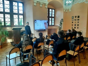 policjantka stoi przy ekranie telewizora, młodzież siedzi na krzesłach tyłem