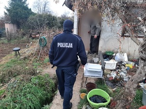 policjant umundurowany zwrócony do obiektywu plecami idzie  w kierunku altany w której drzwiach stoi mężczyzna, wkoło altany nieład