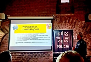 baner z napisem współpraca z samorządami i stojący umundurowany policjant na tle ściany z cegły