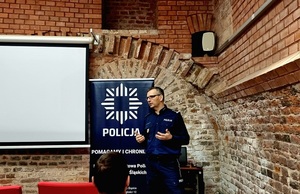 umundurowany policjant stojący na tle ściany z cegły na tle granatowego baneru z gwiazdę policyjną