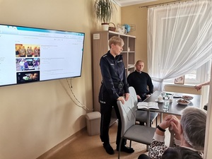 1 umundurowany policjant stoi przy krześle, drugi siedzi przy stole, w pomieszczeniu, za nimi telewizor