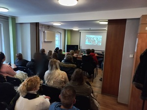 osoby siedzące tyłem na krzesłach w pomieszczeniu, przed nimi ekran telewizora