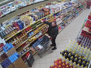 ubrany na ciemno mężczyzna stoi pomiędzy pólkami sklepowymi i sięga po coś z półki