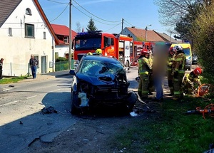 droga, budynki, uszkodzony samochód koloru ciemnego, w tle strażacy