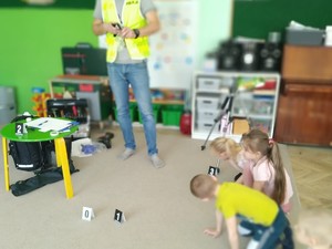 widać postać w kamizelce i plakietki z numerkami na podłodze, dzieci siedzą w pomieszczeniu