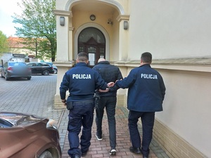 dwóch umundurowanych policjantów prowadzi mężczyznę ubranego na ciemno w kajdankach założonych na ręce trzymane z tylu. Dzień, miejsce zdjęcia przed budynkiem, w tle samochody osobowe
