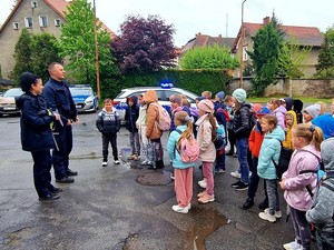 dwóch umundurowanych policjantów stoi obok radiowozu przed dziećmi w wieku szkolnym, światło dzienne, plac, mokra nawierzchnia