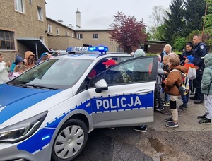 dwóch umundurowanych policjantów stoi obok radiowozu, dzieci w wieku szkolnym stoją obok radiowozu, część wchodzi do środka, światło dzienne, plac, mokra nawierzchnia