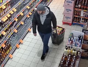 mężczyzna w szarej czapce, ubrany na ciemno, pod kurtą szara bluza z kapturem na terenie sklepu spożywczego