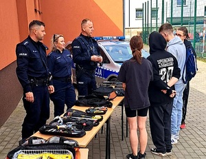 policjanci przy stołach, w tle radiowóz, przed umundurowanymi policjantami stoi młodzież