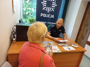 umundurowany policjant siedzi za biurkiem w pomieszczeniu, przed nim na blacie ulotki i kobieta odwrócona tyłem siedząca