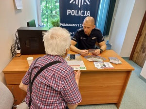 umundurowany policjant siedzi za biurkiem w pomieszczeniu, przed nim na blacie ulotki i mężczyzna odwrócony tyłem siedząca
