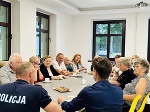 przy jasnym stole siedzą osoby, kobiety i mężczyźni oraz umundurowany policjant, pomieszczenie