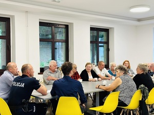 przy jasnym stole siedzą osoby, kobiety i mężczyźni oraz umundurowany policjant, pomieszczenie