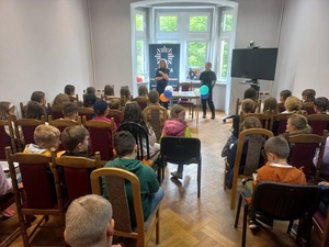 policjantka stoi przy oknie w pomieszczeniu, młodzież siedzi na krzesłach tyłem