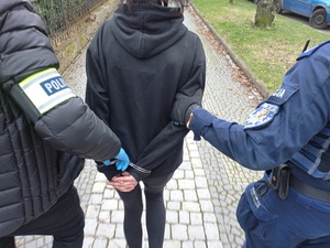 zatrzymana kobieta w kajdankach z tyłu prowadzona przez 2 policjantów chodnikiem, jeden nieumundurowany