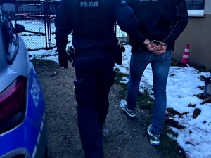 mężczyzna w kajdankach zapiętych z tyłu, prowadzony przez umundurowanego funkcjonariusza