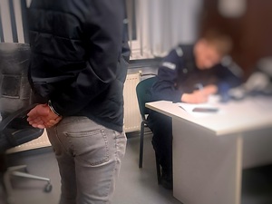 zatrzymany w kajdankach stoi, przy biurku umundurowany policjant sporządza dokumentację